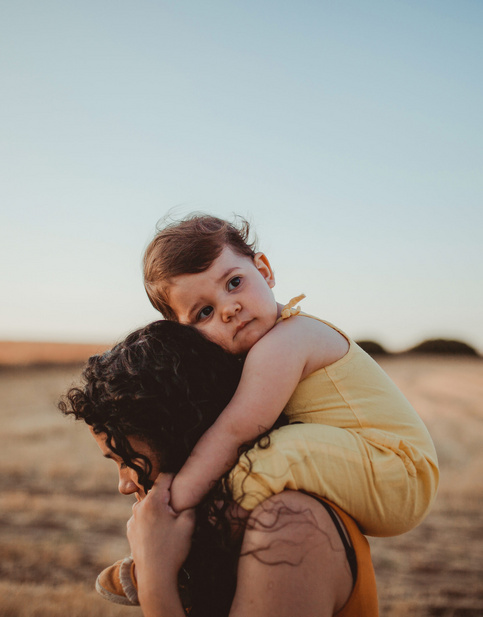 Kleinkind in gelbem Outfit auf den Schultern der Mutter, in einem weiten Feld bei Sonnenuntergang.